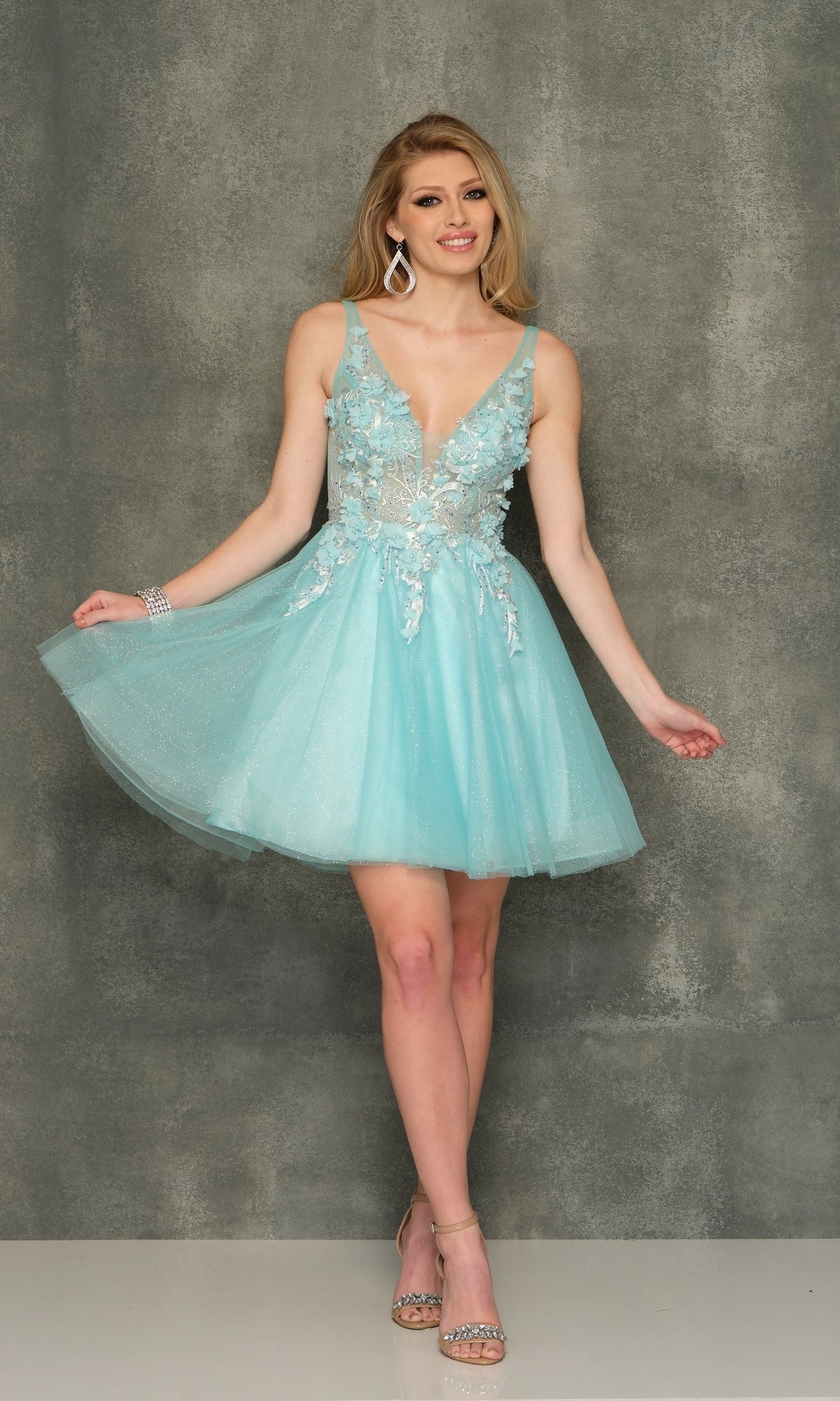 A woman poses against a gray backdrop in the Sheer-Bodice Short Homecoming Dress—a sleeveless, light blue, knee-length party dress with floral embroidery—paired with silver heels, statement earrings, and a bracelet.